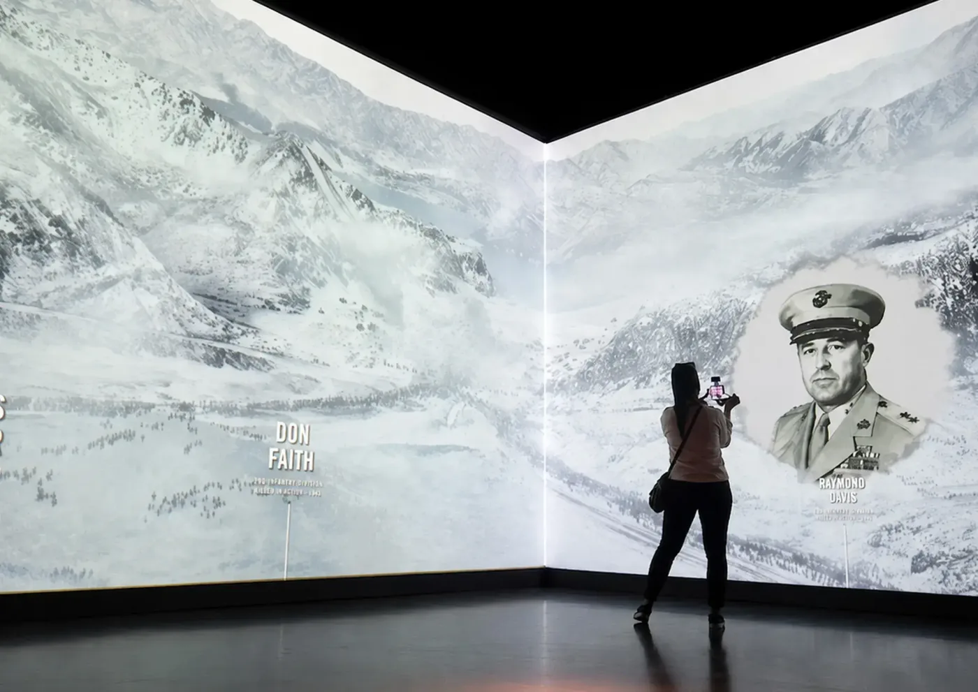 A visitor photographs a floor-to-ceiling projection of a snowy mountain battlefield with a recipient's portrait